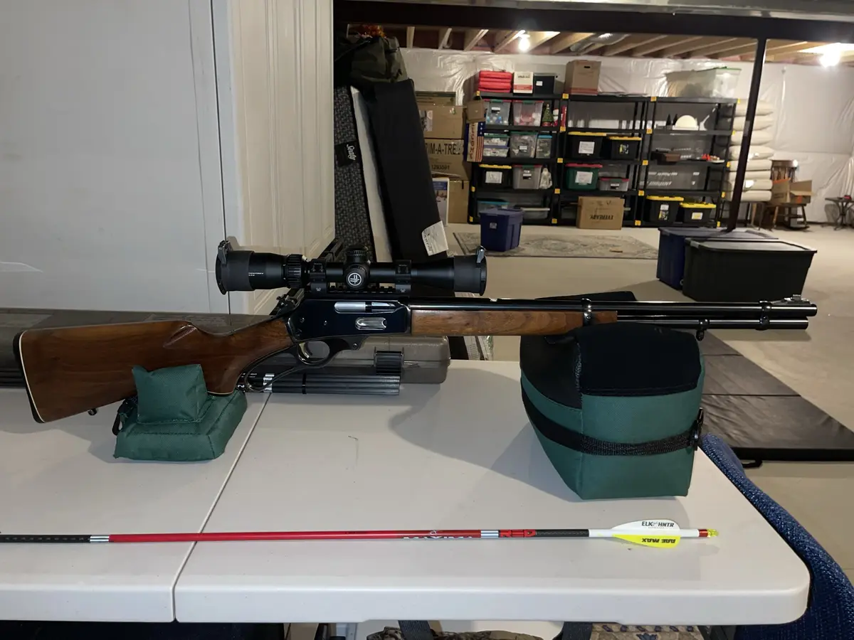 A rifle with a scope rests on a table with green shooting rests, alongside a single arrow with a yellow fletching and red…