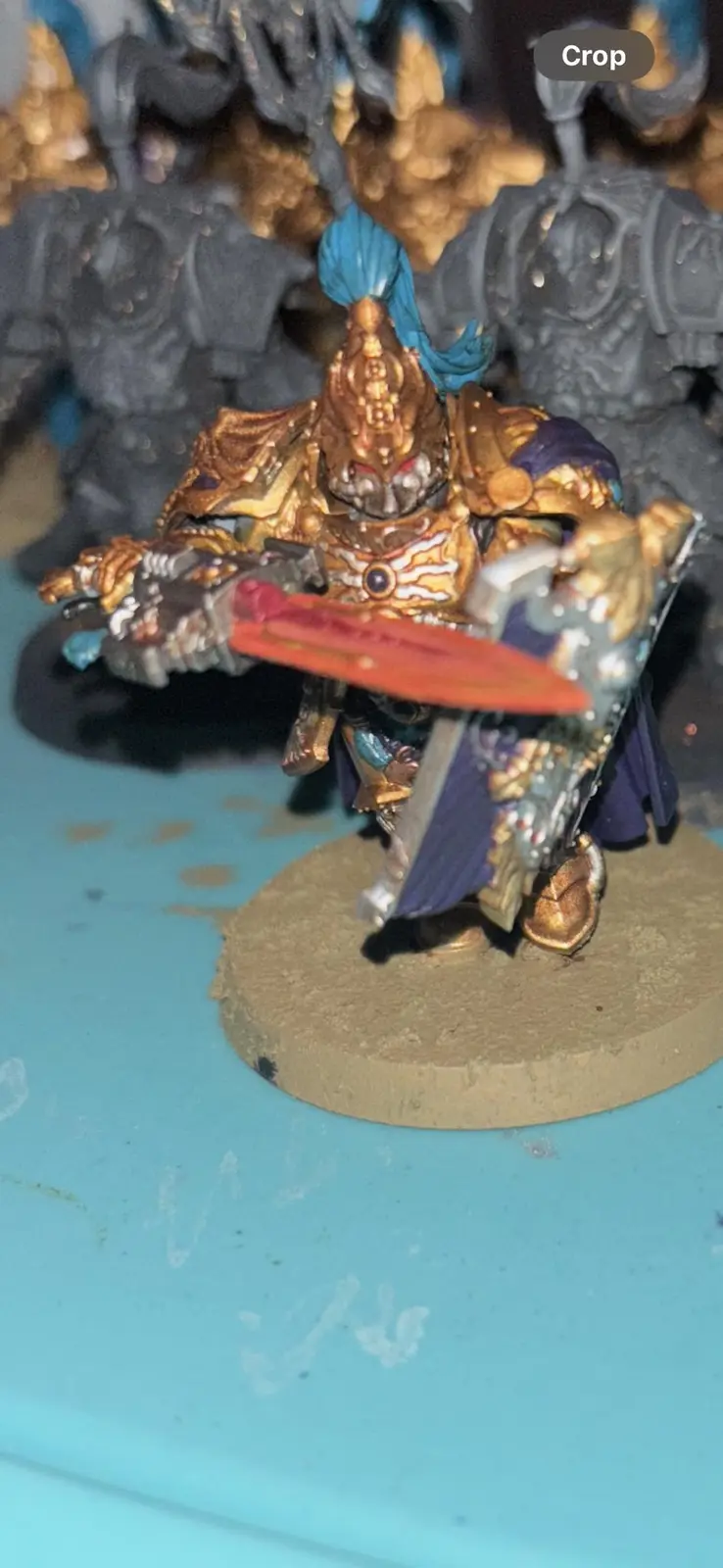 A close-up photo of a painted miniature figure, likely from Warhammer 40,000, wearing ornate golden armor with blue hair and…