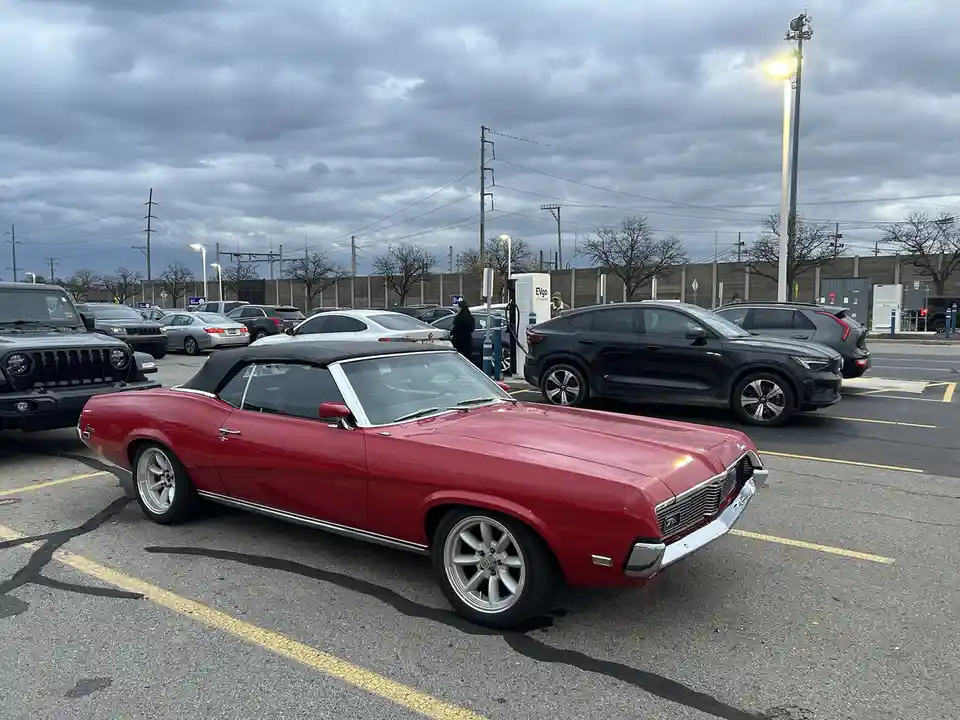 A red classic convertible muscle car is parked in a modern parking lot with other cars, including electric vehicles at…