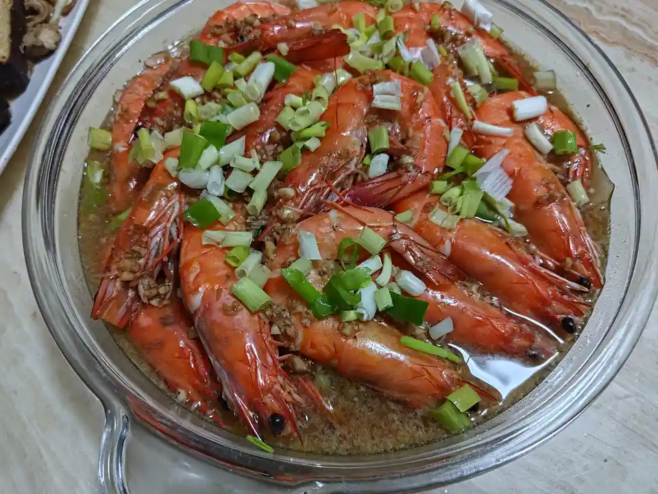 A clear glass bowl filled with large, cooked, bright orange shrimp in a savory sauce, generously topped with chopped green…