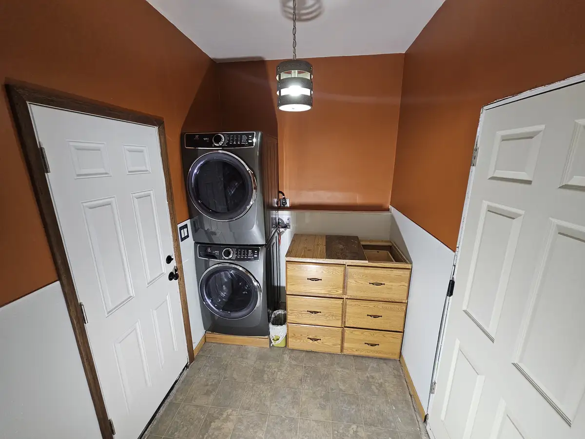 A narrow laundry room with terracotta-colored upper walls and white lower walls, featuring a stacked washer and dryer unit…