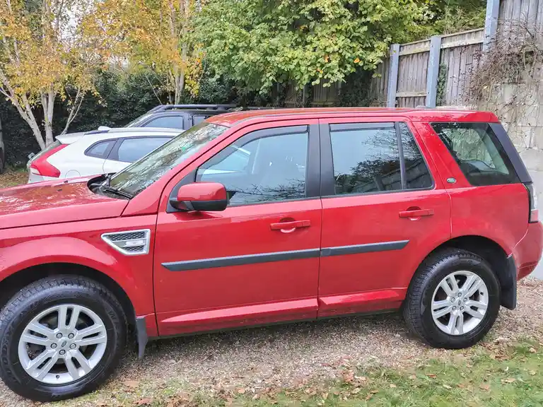 A red Land Rover Freelander 2 SUV is parked on gravel with autumn leaves, showing its side profile. Behind it are other cars…
