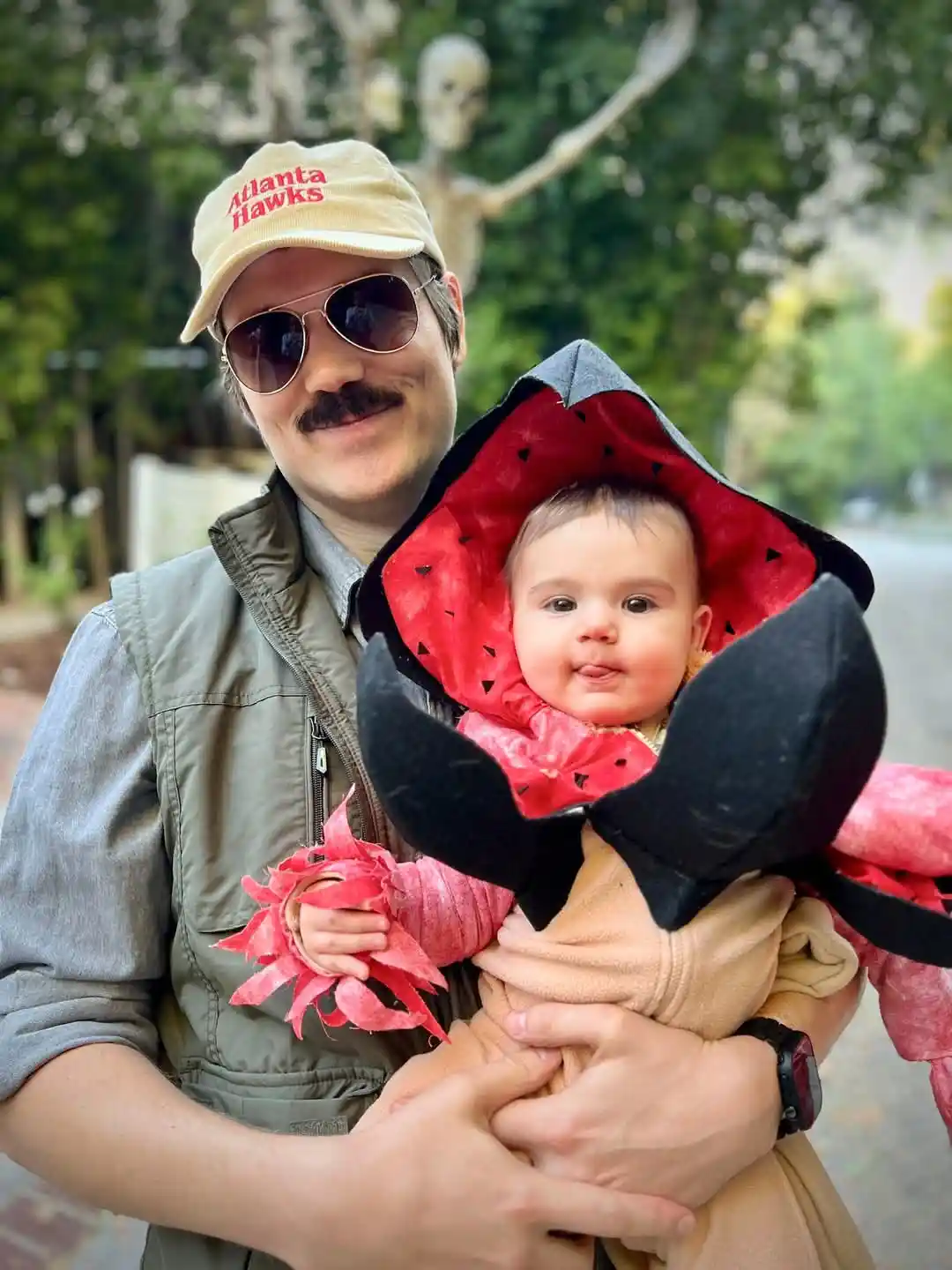 A man wearing a beige 'Atlanta Hawks' cap and sunglasses holds a baby dressed in a watermelon-themed costume with a red hood…