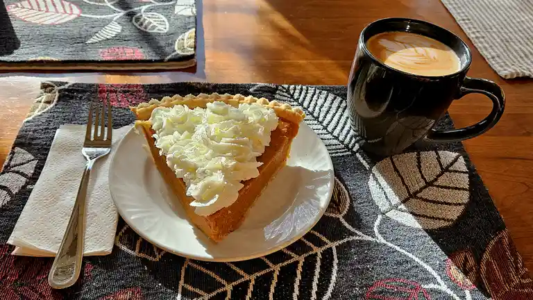 A slice of pumpkin pie topped with whipped cream sits on a white plate next to a black mug of coffee with latte art, both…