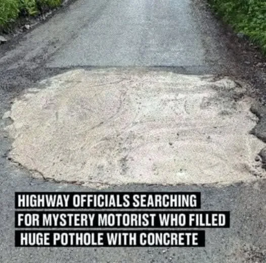 A large, flat, light-gray patch of concrete fills a pothole in the middle of a paved road, surrounded by asphalt and…