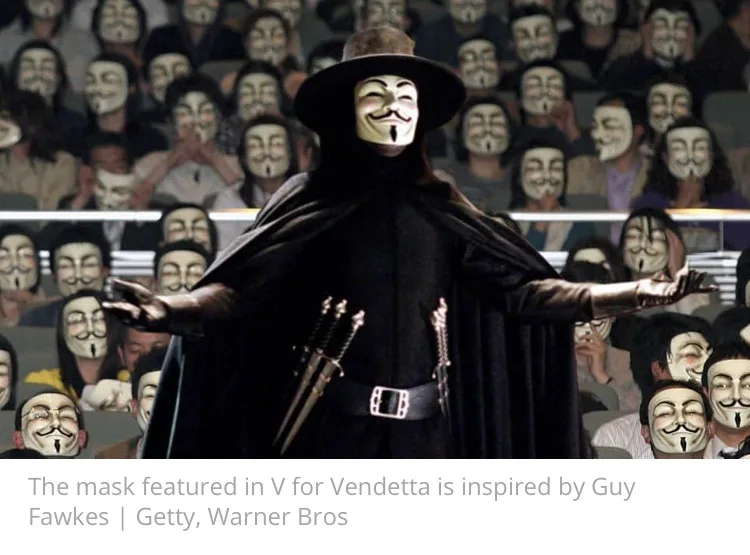 A person in a black cloak and wide-brimmed hat stands center stage with arms outstretched, wearing a white Guy Fawkes mask…