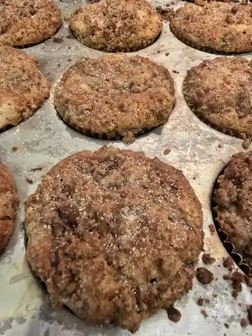A close-up photo shows several golden-brown cinnamon sugar muffins in a metal baking tray, with one muffin in the foreground…
