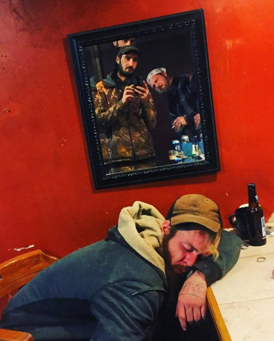 A man with a beard and cap is asleep at a table with a beer bottle nearby, his head resting on his arm. Above him, a framed…