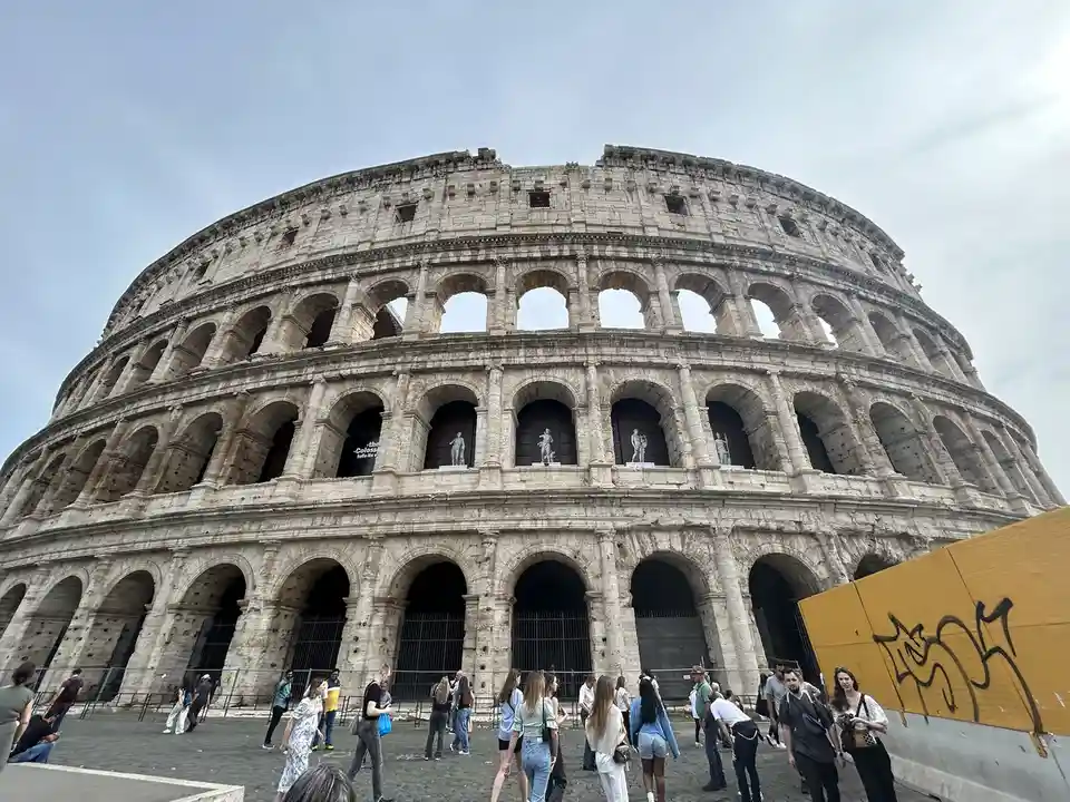 A low-angle photograph shows the massive, ancient Colosseum in Rome, with its iconic stone arches and weathered facade…