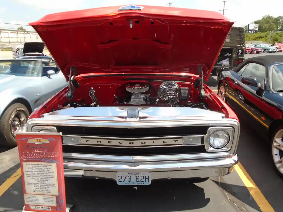 A red Chevrolet pickup truck with its hood open, revealing a shiny engine, is parked at a car show. A red sign in front of…