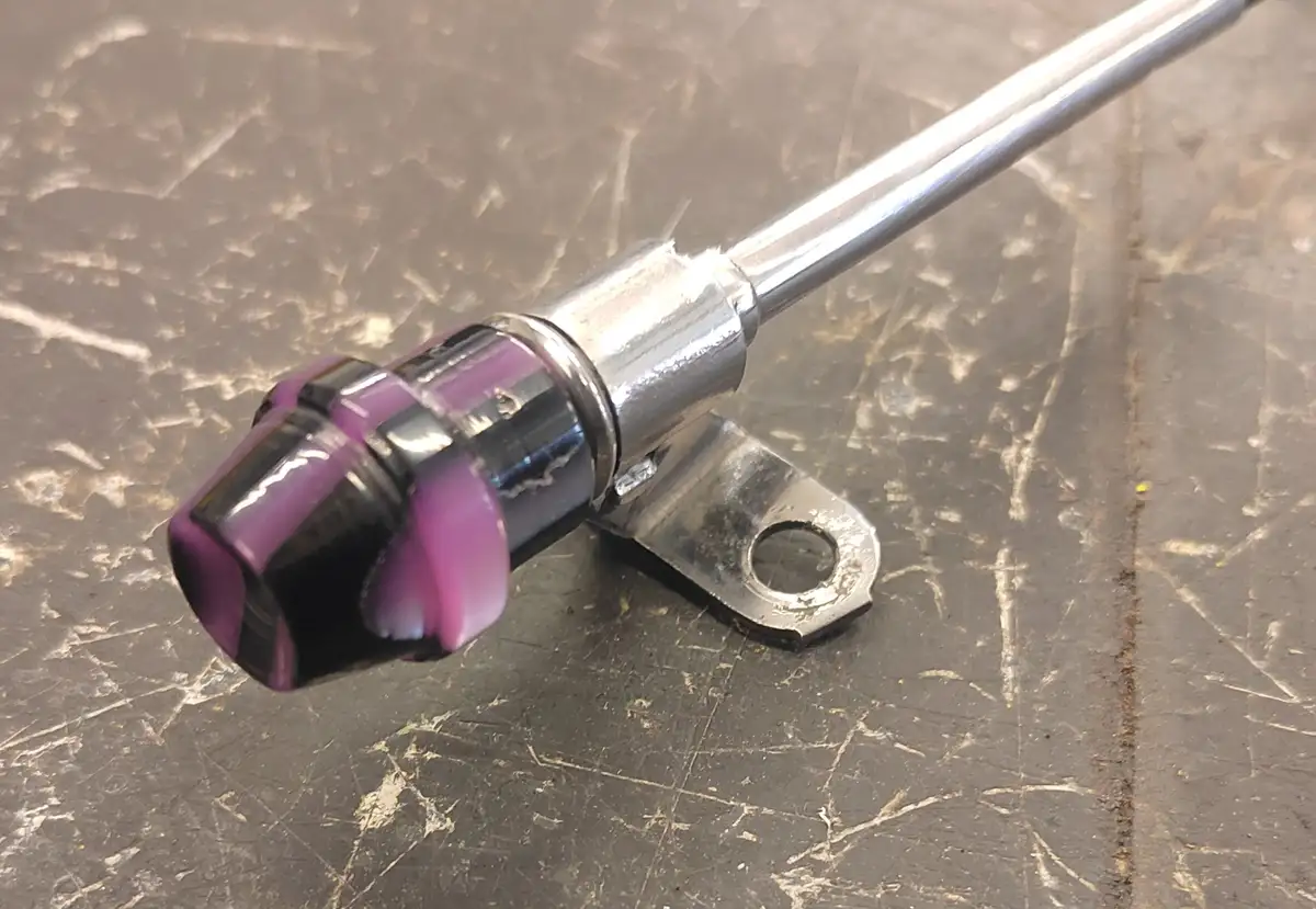 A close-up photo shows a bicycle gear shifter with a shiny chrome shaft and a purple and black handle, mounted on a metal…