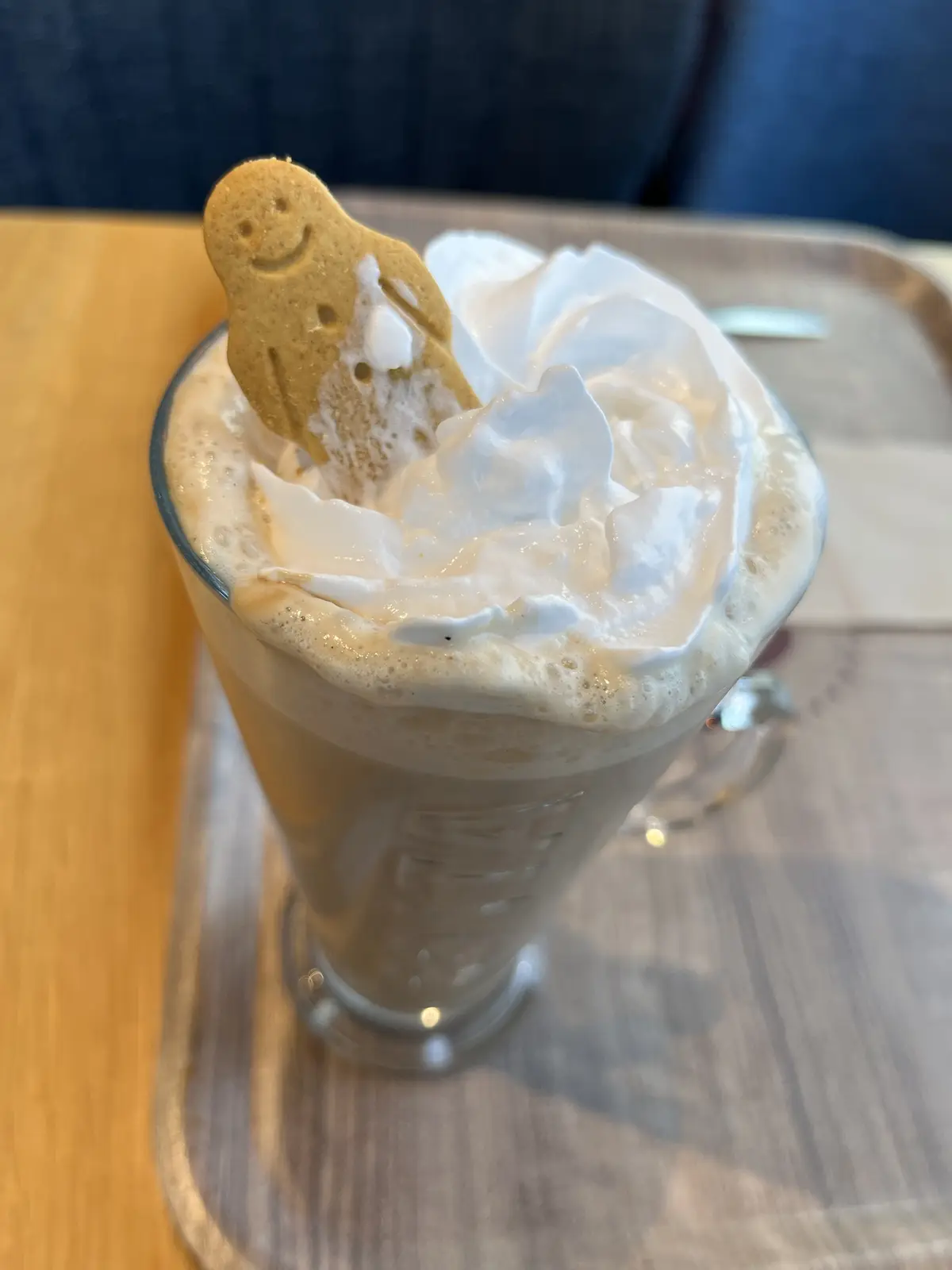 A tall glass of latte topped with whipped cream sits on a wooden tray, with a smiling gingerbread man cookie stuck into the…