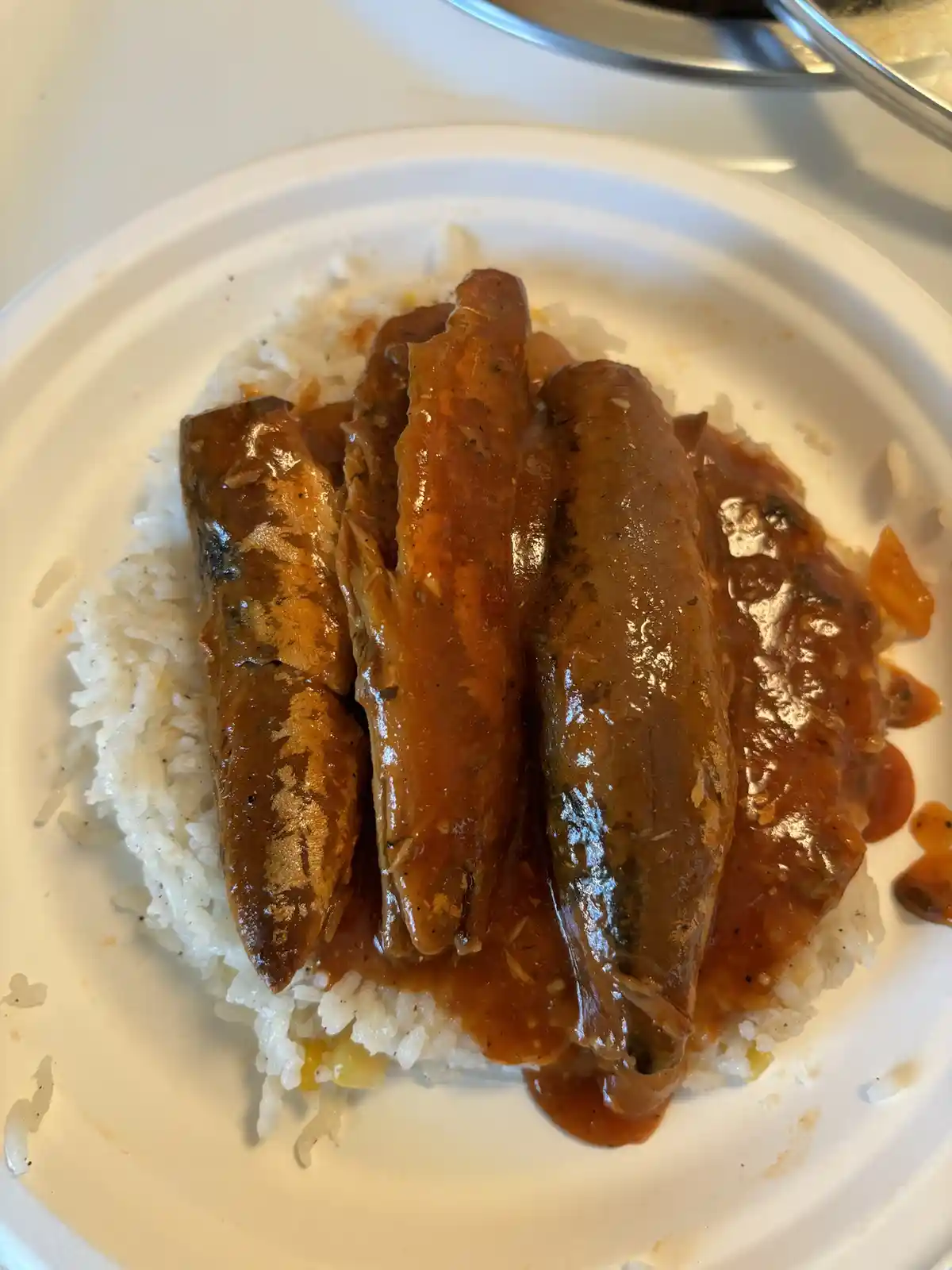 A close-up photo shows several whole sardines covered in a thick, reddish-brown sauce, served atop a bed of white rice on a…