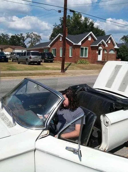 A person with curly hair sits in the driver's seat of a white convertible with the top down and hood open, looking tired or…