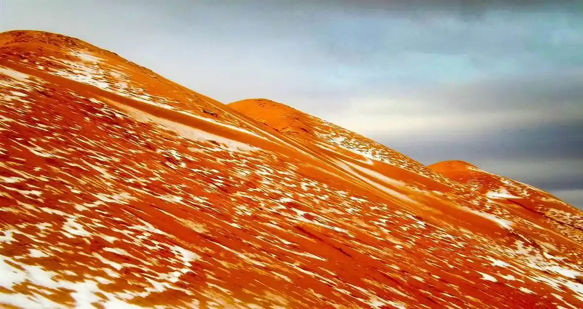 A wide shot of reddish-orange sand dunes partially covered with white snow under a cloudy sky, with a small dark vehicle…