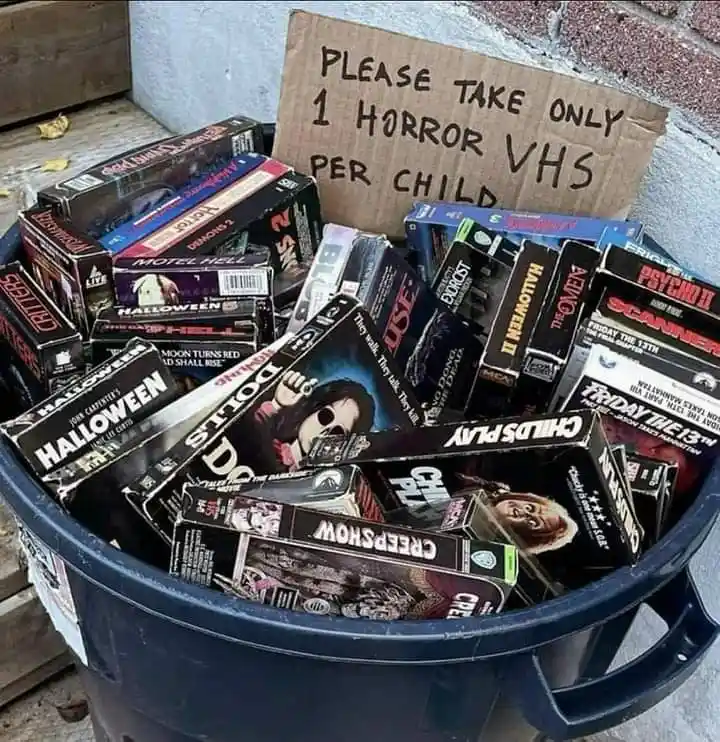 A blue trash bin overflowing with vintage horror VHS tapes, including titles like 'Halloween', 'Child's Play', and 'Friday…