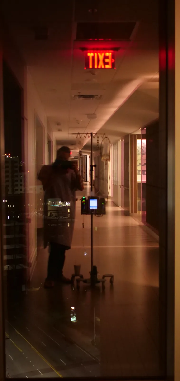 A blurry reflection in a glass door shows a person in a hospital gown holding a phone, standing beside an IV pole with a bag…