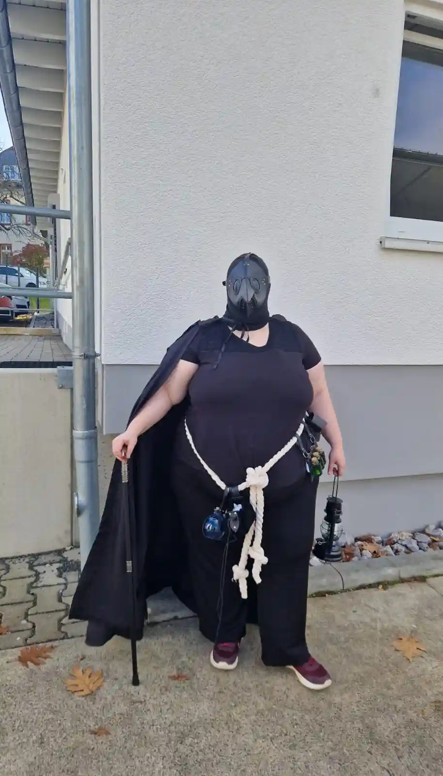A person wearing a black plague doctor mask, a black cape, and black clothing stands outside a building holding a cane and a…