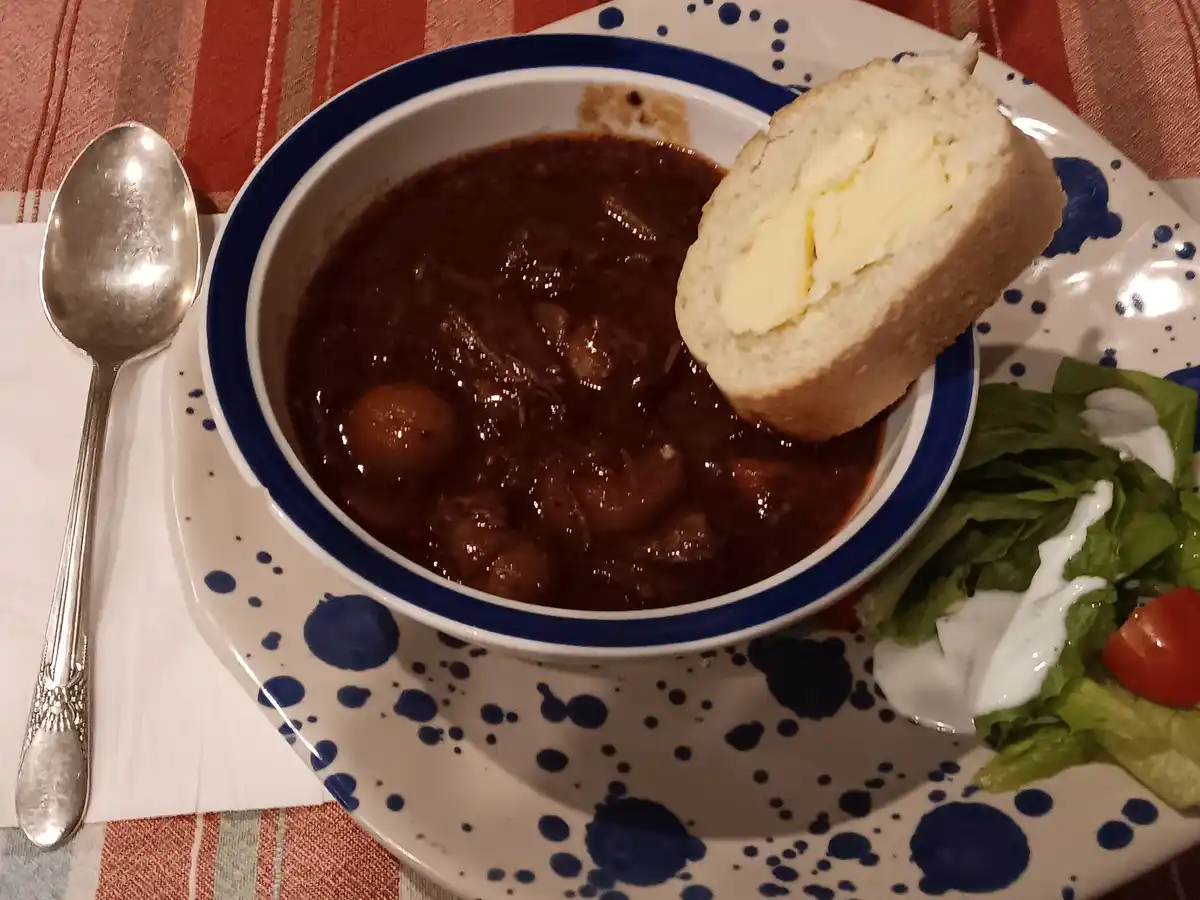 A bowl of dark, thick stew with chunks of meat and vegetables sits on a blue-speckled plate, accompanied by a buttered bread…