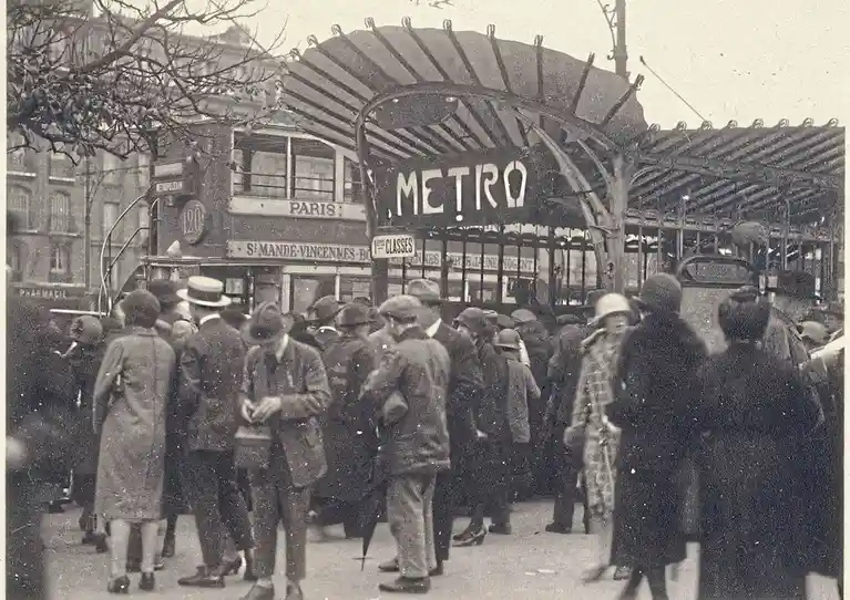 A vintage black-and-white photograph shows a crowded Parisian street scene with people gathered near a large, ornate metal…