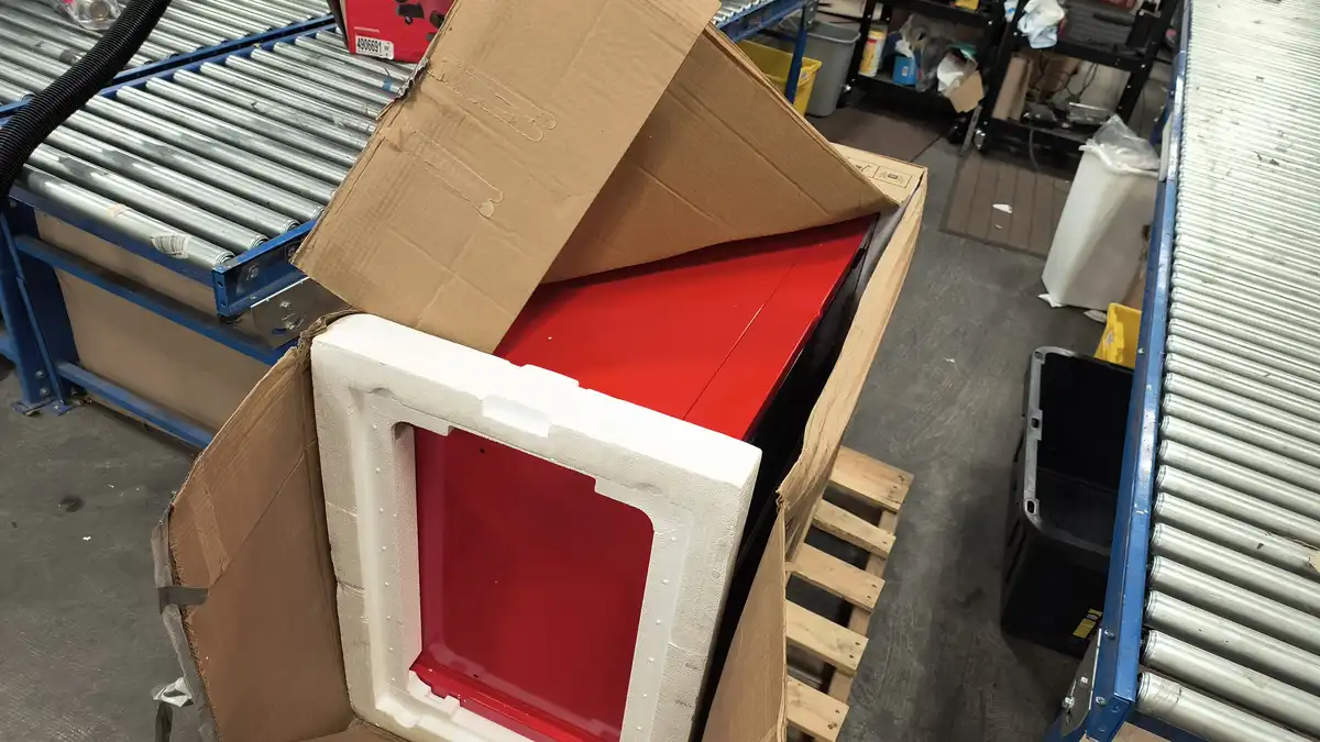 A red appliance, likely a fridge, is partially unpacked from a cardboard box in a warehouse setting, surrounded by…