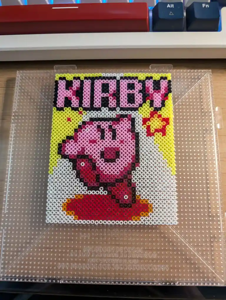 A pixel art portrait of the video game character Kirby made from colorful perler beads on a transparent pegboard, placed on…