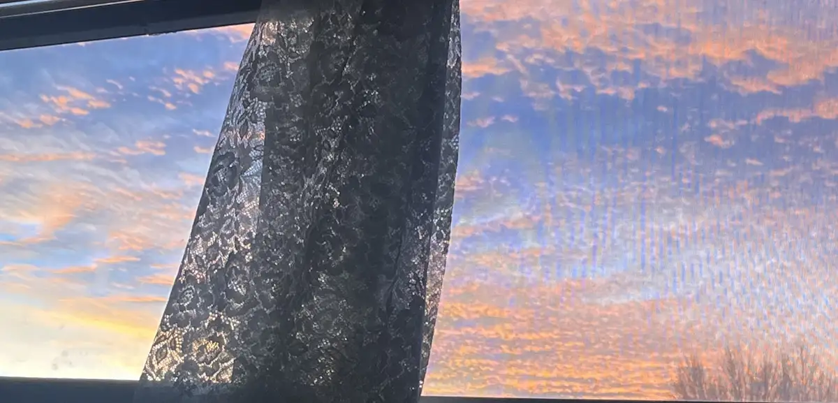 A photo shows a black lace curtain partially covering a window with a vibrant sunset sky of orange and blue clouds visible…