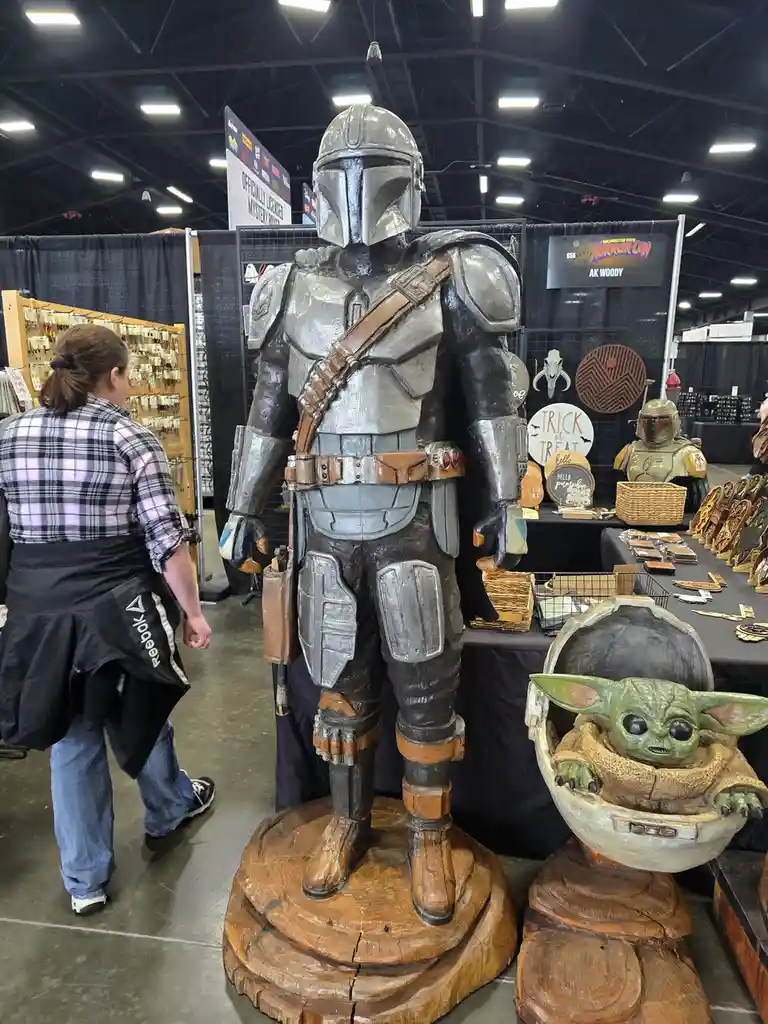 A large, detailed wooden carving of The Mandalorian stands on a tree stump base at a convention, with a smaller carved Baby…