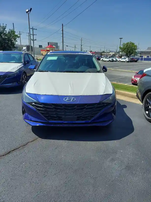 A shiny blue Hyundai car is parked in a dealership lot with a white protective cover draped over its hood and windshield…