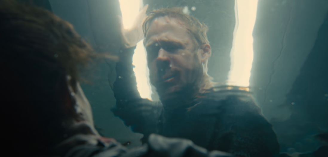 A man with wet hair and a distressed expression is submerged underwater, reaching upward toward a bright light source above…