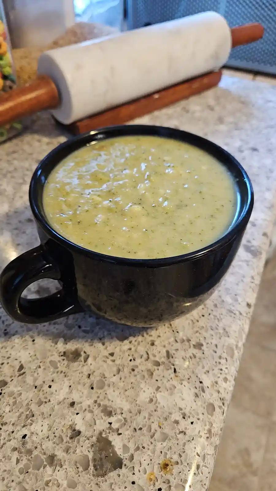 A black mug filled with light green, speckled soup sits on a speckled countertop, with a white marble rolling pin with…