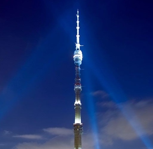 A tall, illuminated tower, likely the Ostankino Tower in Moscow, stands against a dark blue night sky with beams of light…