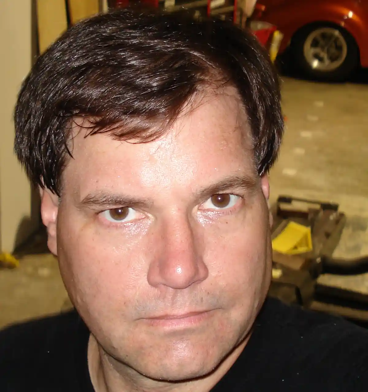A close-up photo of a man with sweaty, glistening skin and a stern, unamused expression, set against a garage background…
