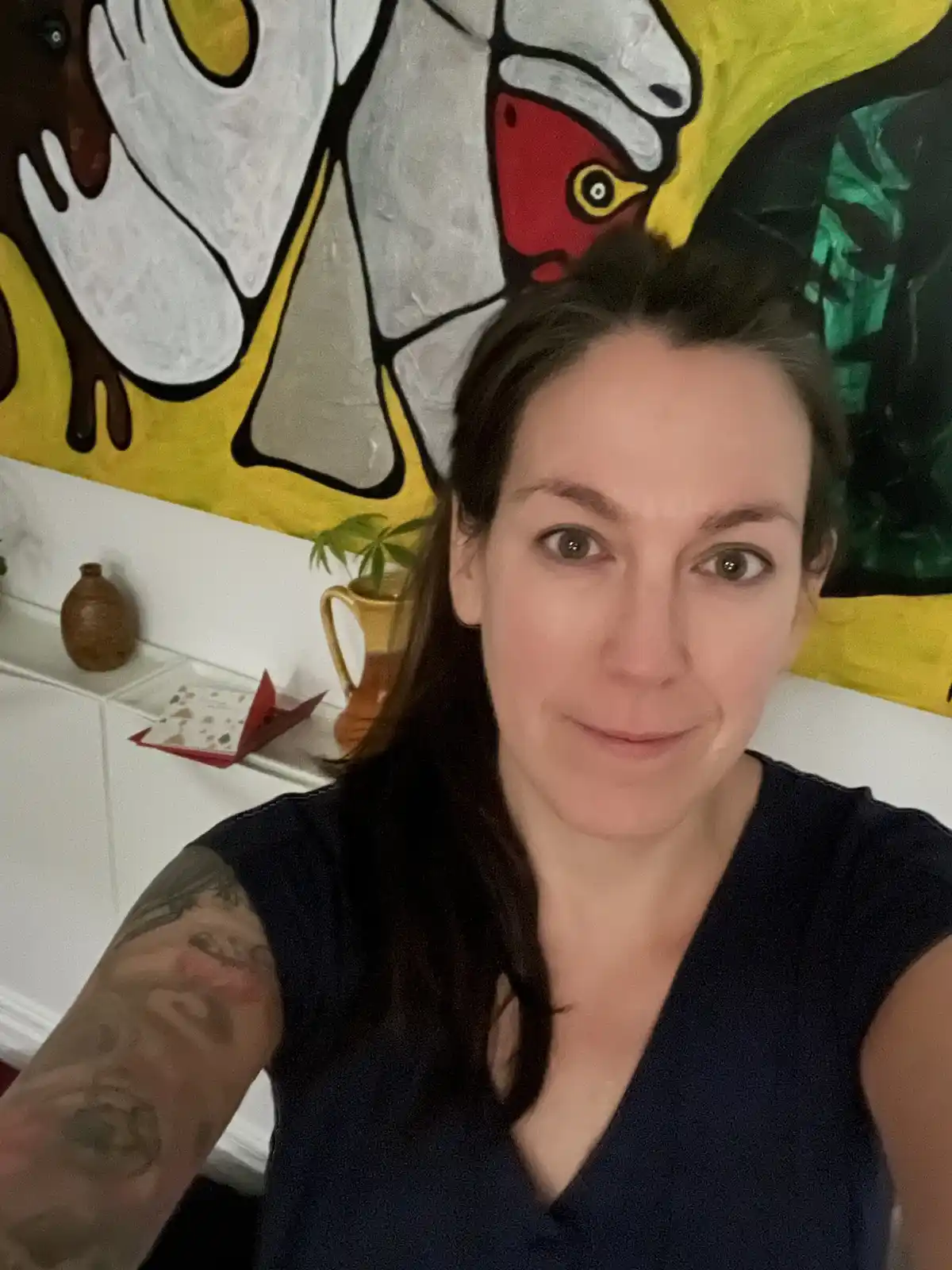 A woman with dark hair and a visible tattoo on her left arm takes a selfie in front of a large, colorful abstract painting…