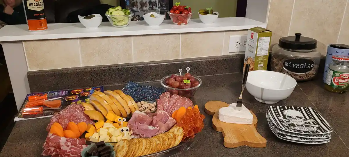 A charcuterie board with meats, cheeses, crackers, and fruit is arranged on a kitchen counter. A wedge of white cheese on a…