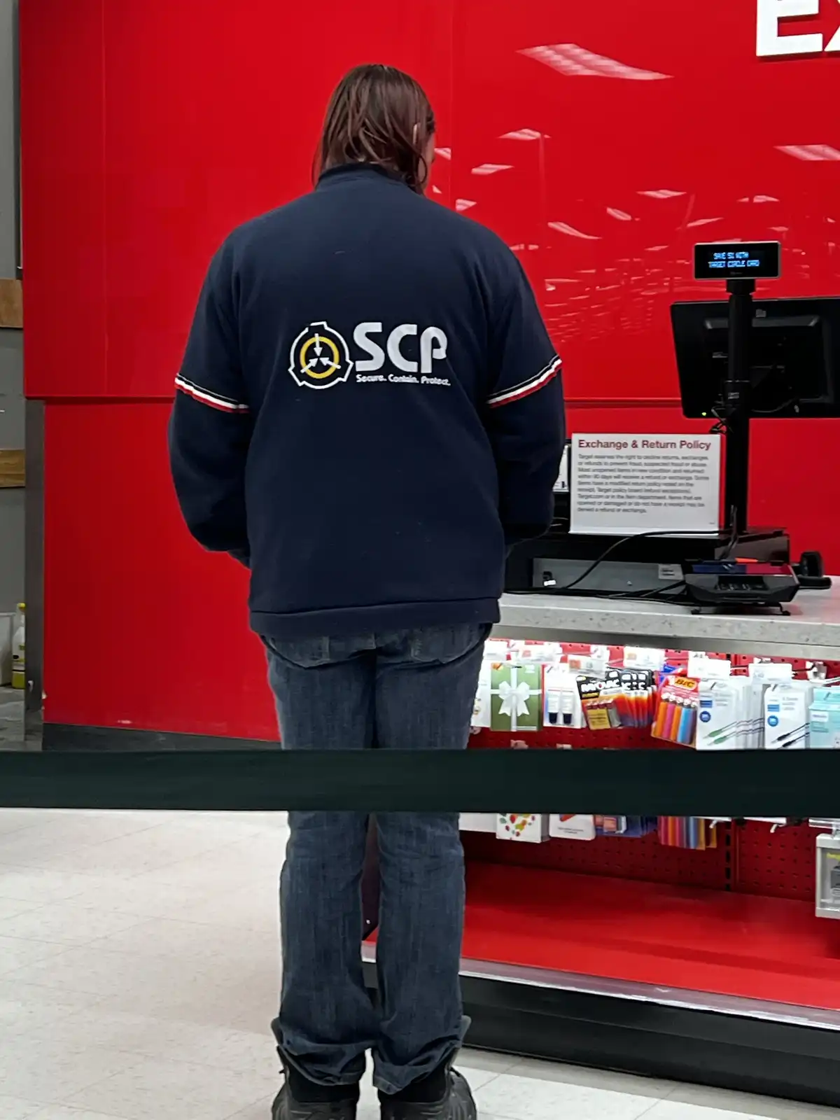 A person with long hair, viewed from behind, wears a dark blue jacket with the SCP Foundation logo and slogan 'Secure…