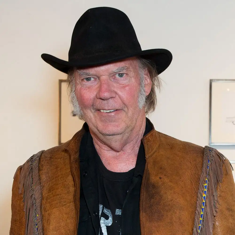 a man with gray hair and a black hat wears a brown fringed leather vest over a black t-shirt. He smiles slightly while…