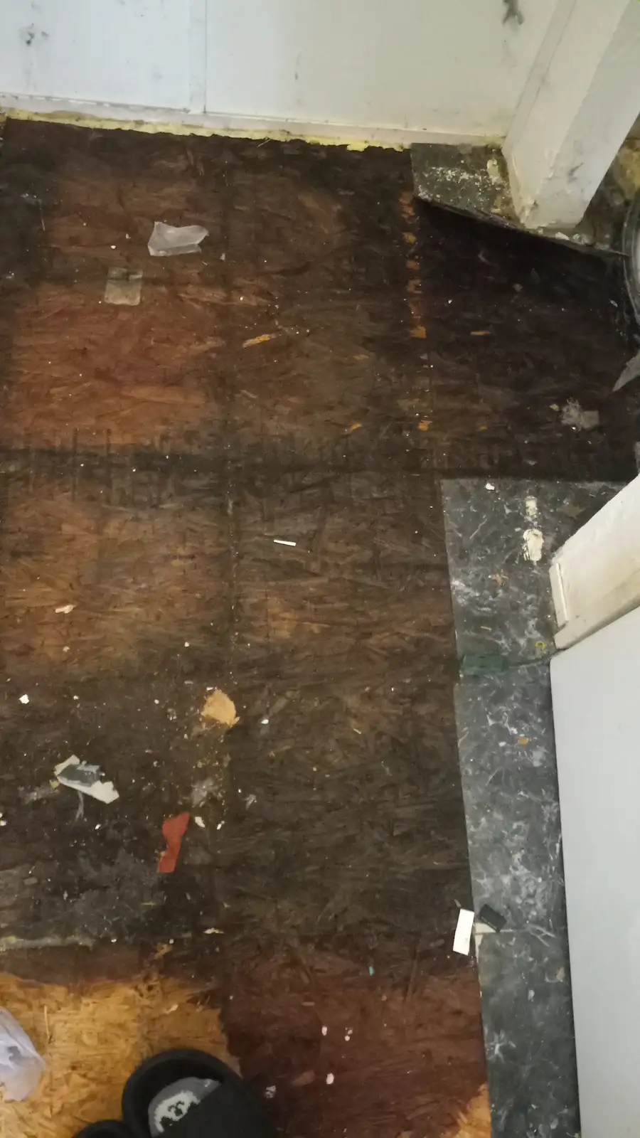 A top-down view of a messy, partially stripped floor showing dark plywood sheets, scattered debris, and a mix of flooring…