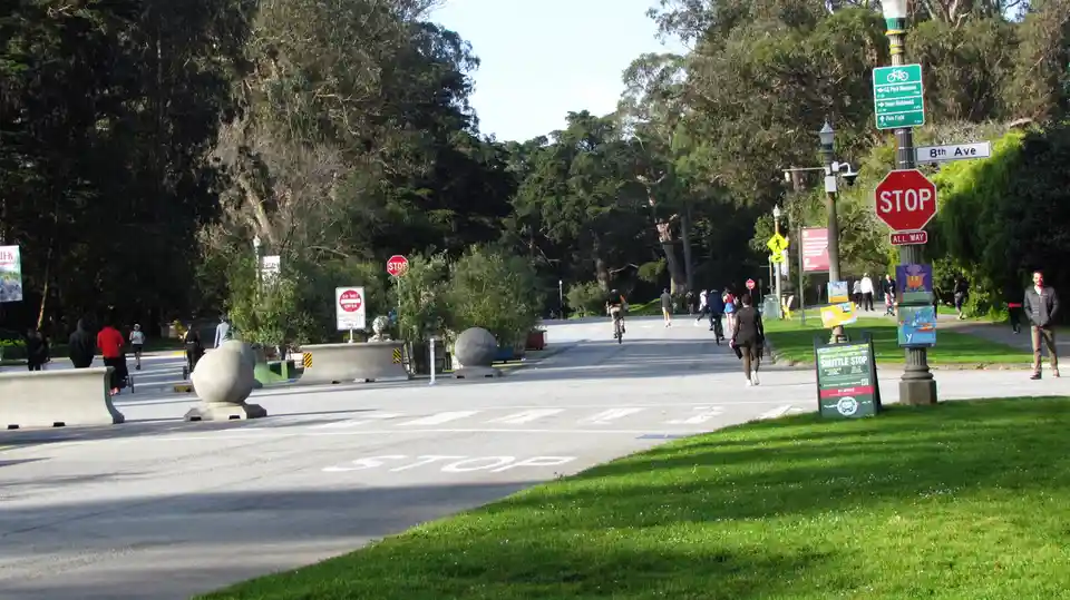 A wide, tree-lined street in a park with pedestrians, cyclists, and concrete barriers. A red stop sign and an 'ALL WAY' sign…