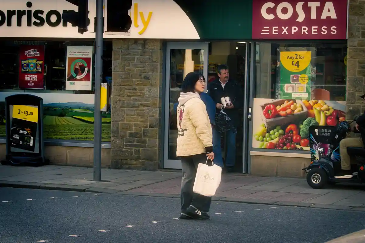A woman in a beige quilted jacket and glasses walks across a street in front of a Morrisons Daily store with a Costa Express…