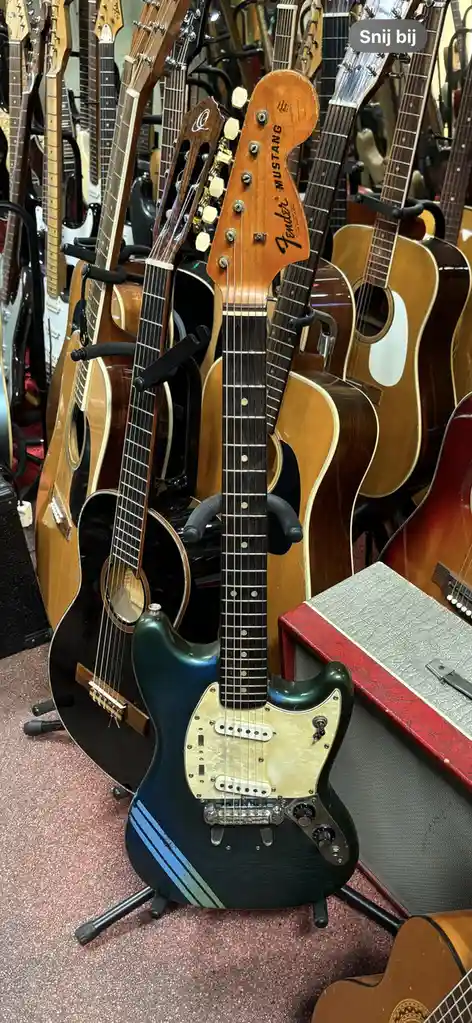 A Fender Mustang electric guitar with a dark green finish and blue stripes stands in a music store surrounded by other…