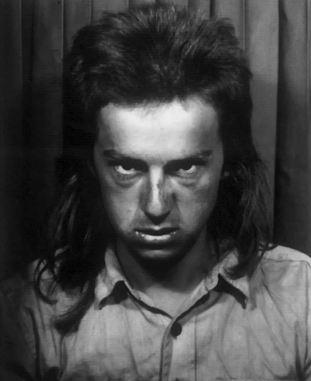 A black-and-white mugshot of a man with long, unkempt hair and a weary, intense expression, wearing a simple collared shirt…