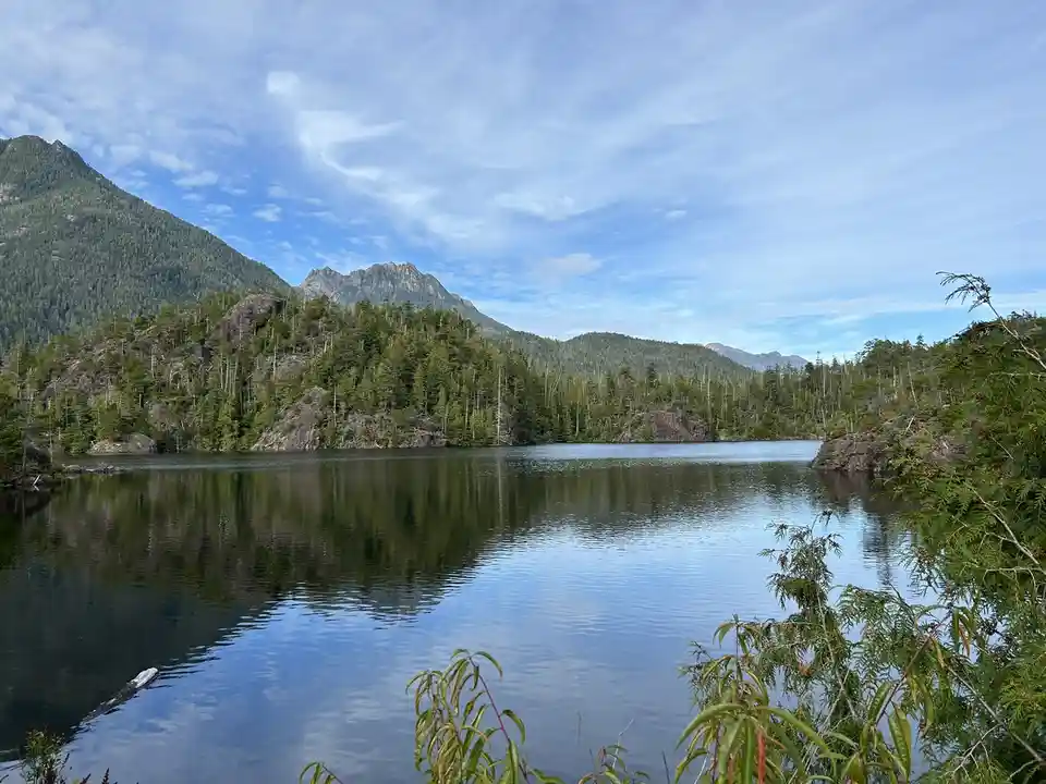 A serene landscape photo of a calm lake surrounded by forested mountains under a partly cloudy sky. The image has text…