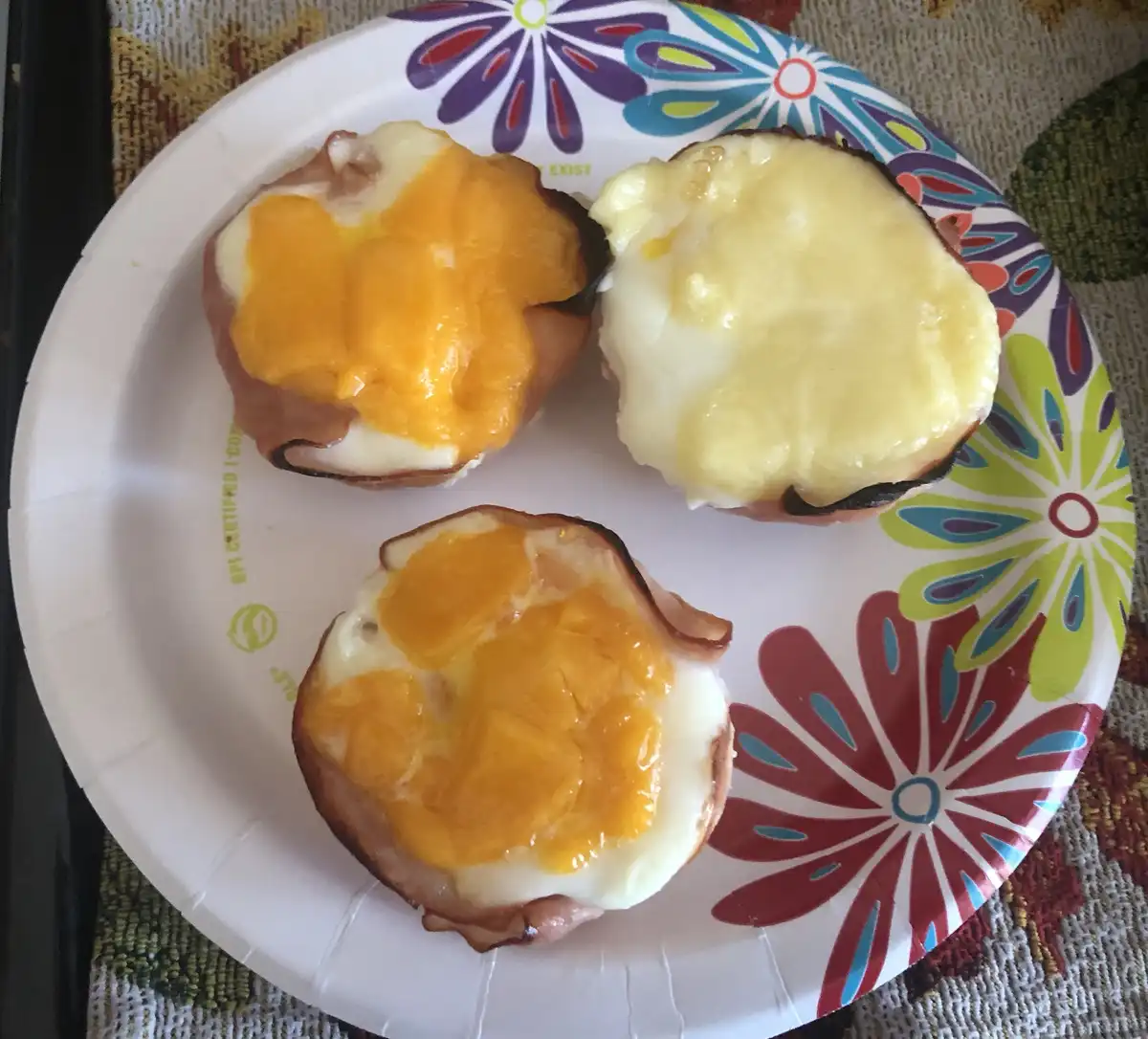 Three baked egg and cheese breakfast cups sit on a colorful floral paper plate, each with a different cheese melt—orange…