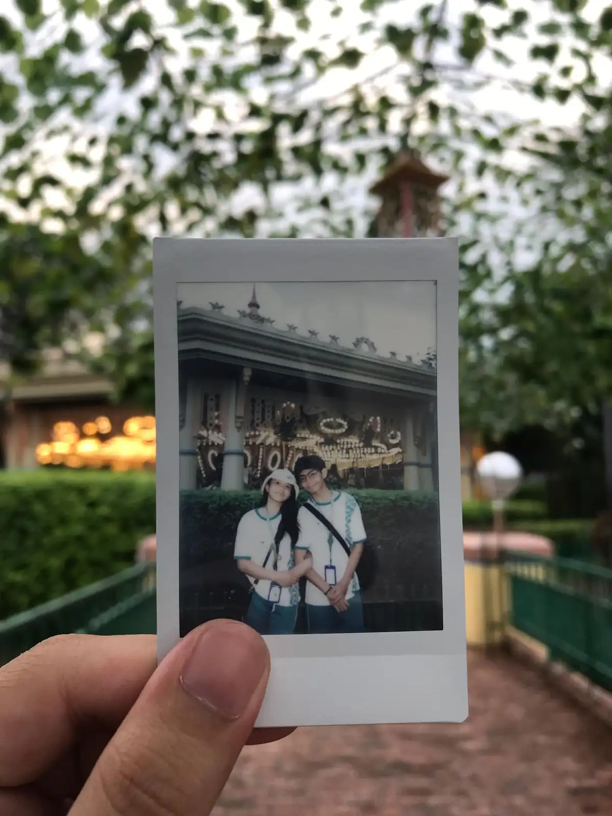 A hand holds up a Polaroid photo of two smiling young people posing in front of a decorative building. The background is…