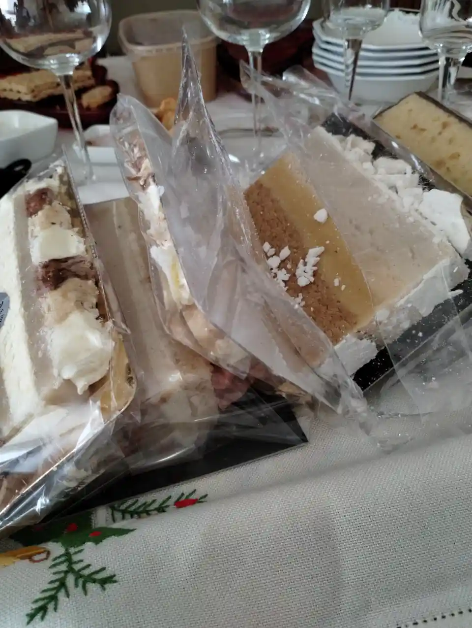 Several individually wrapped slices of cake sit on a tablecloth with a Christmas tree pattern, surrounded by wine glasses…