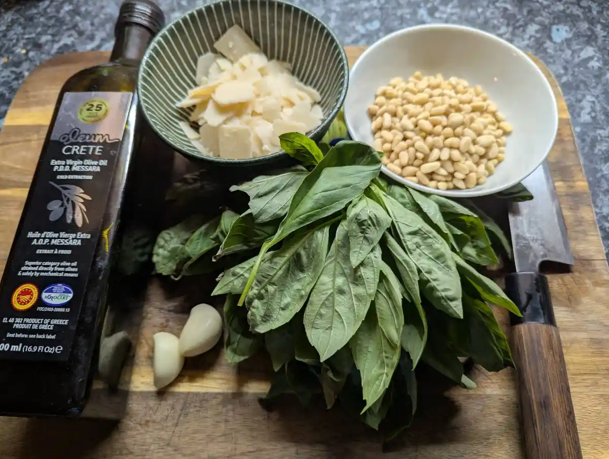 A wooden cutting board displays ingredients for cooking: a bottle of olive oil, bowls of pine nuts and shaved cheese, a…