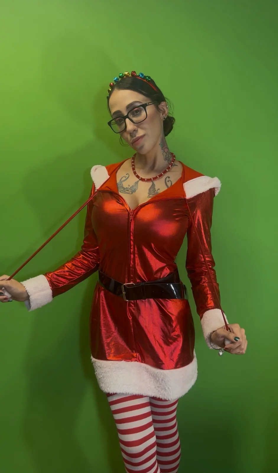 A woman in a shiny red Santa outfit with white trim, red and white striped tights, and a colorful bell headband stands…