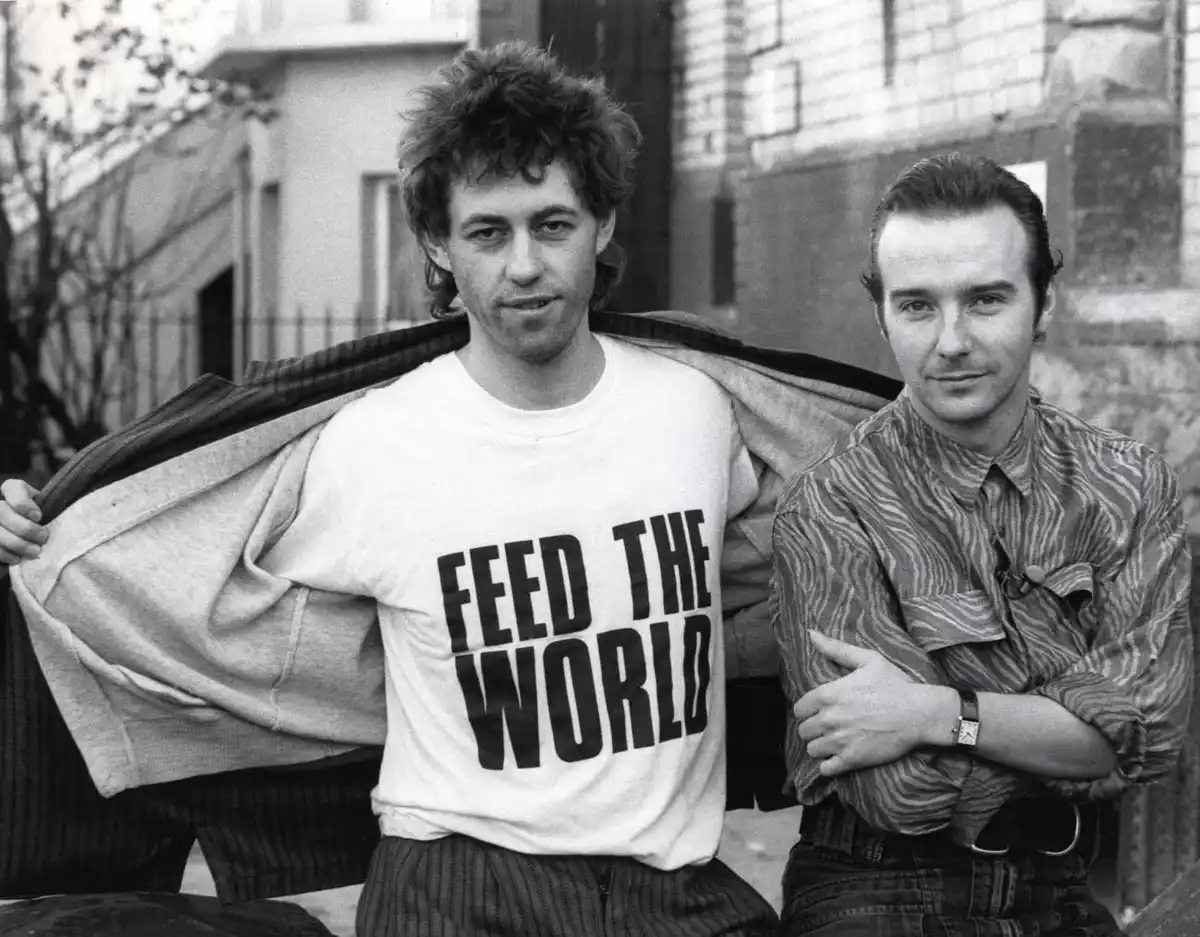 A black-and-white photo shows two men posing outdoors. The man on the left wears a white t-shirt with the bold text 'FEED…