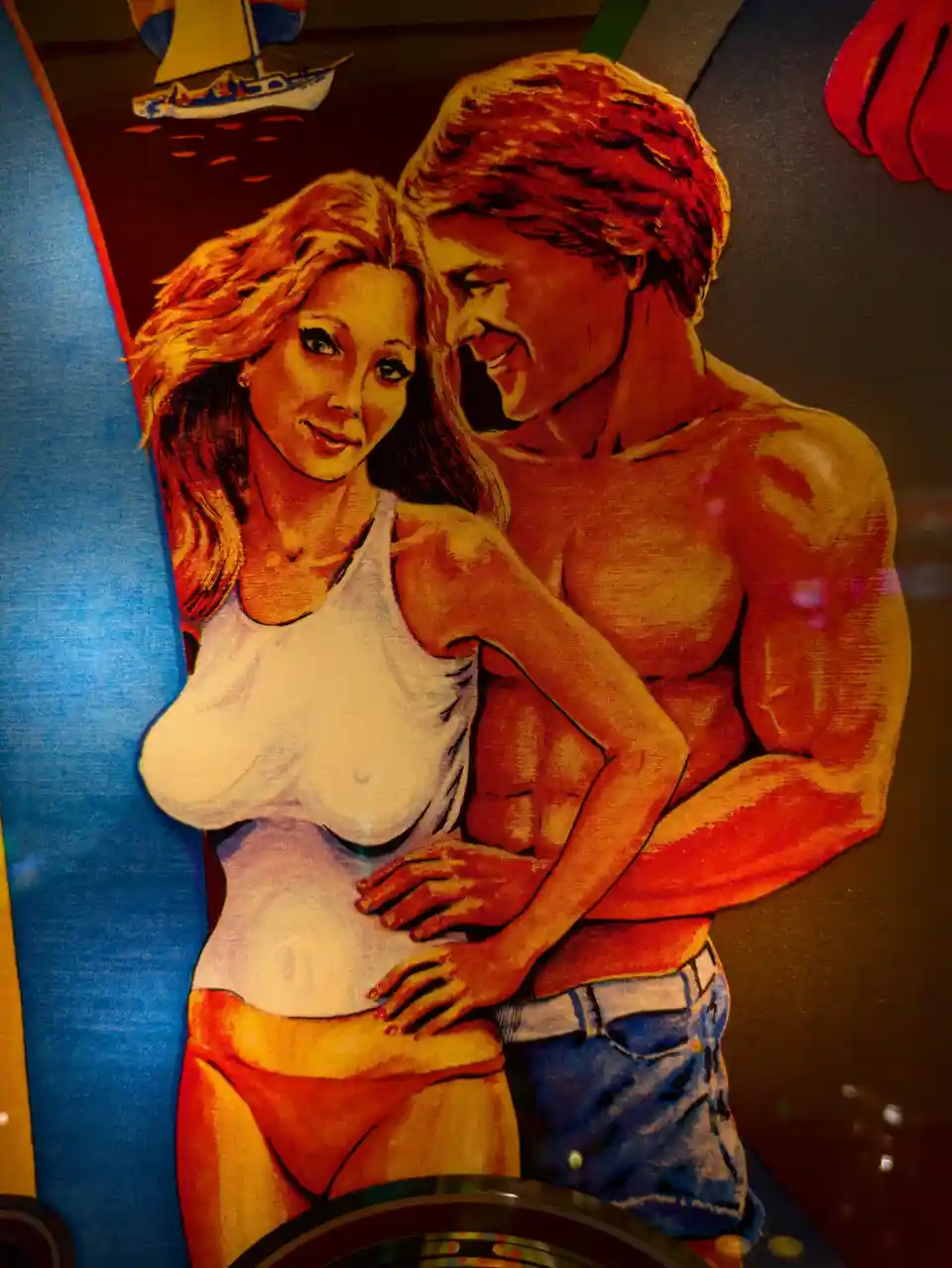 A close-up of a vintage-style pinball machine artwork showing a muscular man in blue shorts hugging a woman in a white tank…